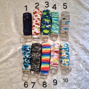 May include: Ten fabric-covered wristlet keychains arranged in two rows, numbered one through ten. Each features a silver-tone metal clasp and a circular split ring. The patterns include floral, geometric, rainbow, spiderweb, and abstract designs.