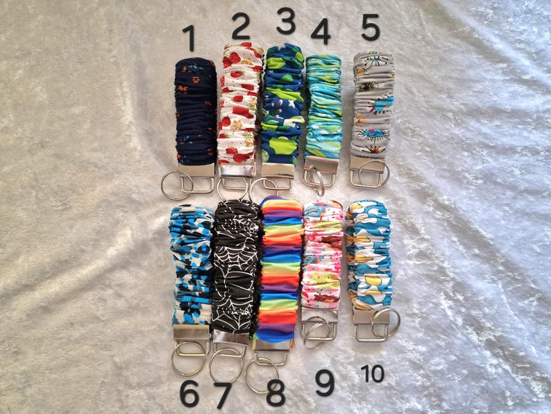 May include: Ten fabric-covered wristlet keychains arranged in two rows, numbered one through ten. Each features a silver-tone metal clasp and a circular split ring. The patterns include floral, geometric, rainbow, spiderweb, and abstract designs.