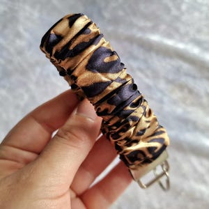 May include: A fabric keychain wristlet featuring a gathered, scrunchie-style design with a black and gold leopard print pattern. The cylindrical fabric section is attached to a silver-toned metal hardware clasp with a circular ring for keys.