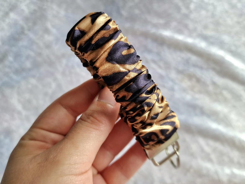 May include: A fabric keychain wristlet featuring a gathered, scrunchie-style design with a black and gold leopard print pattern. The cylindrical fabric section is attached to a silver-toned metal hardware clasp with a circular ring for keys.