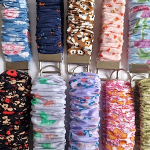 May include: Ten fabric-covered wristlet keychains arranged in two rows. Each keychain features a gathered, elasticized fabric strap with various floral and geometric patterns in colors including blue, black, brown, pink, purple, and green, attached to a silver-tone metal clasp and ring.