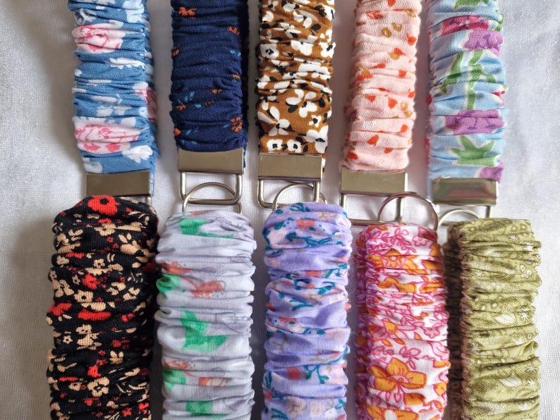 May include: Ten fabric-covered wristlet keychains arranged in two rows. Each keychain features a gathered, elasticized fabric strap with various floral and geometric patterns in colors including blue, black, brown, pink, purple, and green, attached to a silver-tone metal clasp and ring.