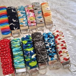 May include: A collection of twelve fabric wristlet keychains featuring various patterns including rainbow stripes, black with white shapes, blue with yellow accents, floral, spiderwebs, strawberries, and red with white stars, each with a metal clasp.