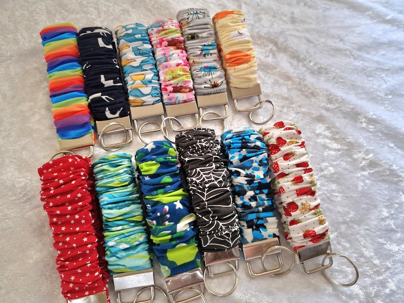 May include: A collection of twelve fabric wristlet keychains featuring various patterns including rainbow stripes, black with white shapes, blue with yellow accents, floral, spiderwebs, strawberries, and red with white stars, each with a metal clasp.