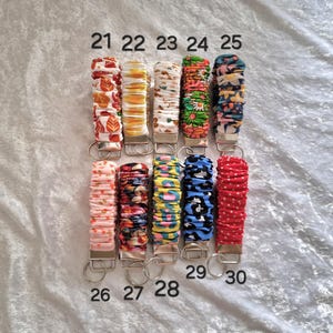 May include: Ten fabric-covered wristlet keychains arranged in two rows, numbered 21 through 30. Each keychain features a gathered, scrunchie-style textile band with various colorful patterns, including floral, animal print, and polka dots, attached to a silver metal clasp.