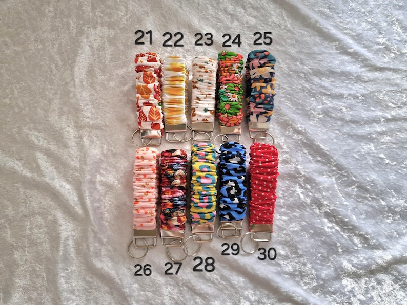 May include: Ten fabric-covered wristlet keychains arranged in two rows, numbered 21 through 30. Each keychain features a gathered, scrunchie-style textile band with various colorful patterns, including floral, animal print, and polka dots, attached to a silver metal clasp.