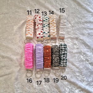 May include: Ten fabric wristlet keychains arranged in two rows, numbered 11 through 20. Each features a gathered, scrunchie-style fabric strap in various colors and patterns, including hearts, polka dots, and gingham, attached to a silver metal clasp.