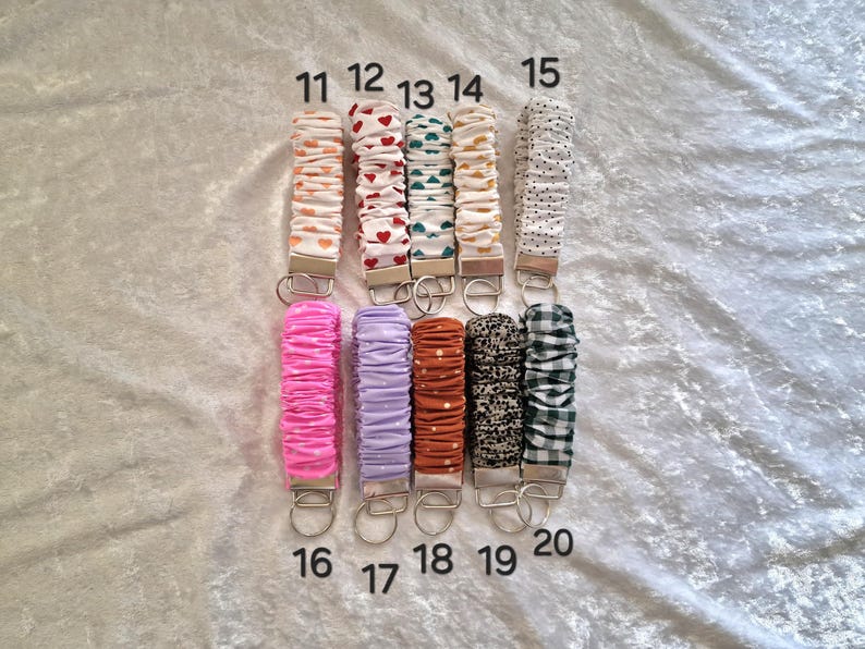 May include: Ten fabric wristlet keychains arranged in two rows, numbered 11 through 20. Each features a gathered, scrunchie-style fabric strap in various colors and patterns, including hearts, polka dots, and gingham, attached to a silver metal clasp.
