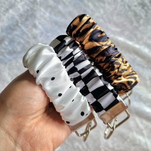 May include: Three fabric-covered keychains featuring a gathered, scrunchie-style design. One is white with black polka dots, one has a black and white checkered pattern, and the third features a brown and black leopard print. Each has a metal ring.