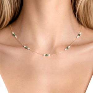 May include: A delicate gold chain necklace featuring small, evenly spaced white rice pearls and tiny green beads. The necklace rests against the neck, showcasing a minimalist design with alternating pearl and green bead accents along the thin metal chain.