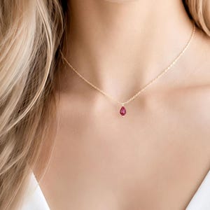 May include: A delicate gold chain necklace featuring a single, small, teardrop-shaped ruby pendant. The necklace rests against the collarbone, showcasing a thin, polished metal link chain and a vibrant, deep red gemstone suspended at the centre.