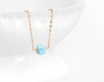 May include: A delicate gold chain necklace featuring a single, small, round, light blue stone bead with white marbled patterns. The necklace is suspended, showcasing the fine metal links and the smooth, polished texture of the central gemstone.