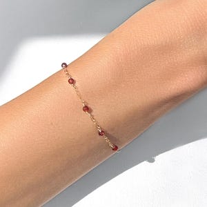 May include: A delicate gold chain bracelet featuring small, round, deep red gemstone beads spaced evenly along the length of the chain. The bracelet is worn around the wrist, showcasing the thin metallic links and the dark red stones.