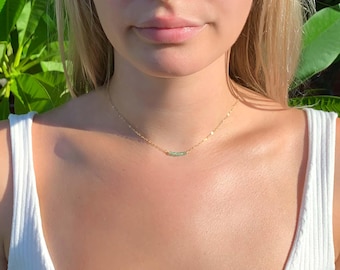 May include: A delicate gold chain necklace featuring a small, horizontal, light green cylindrical bead at the center. The necklace rests against the neck, showcasing a thin metallic link chain and a single, smooth, pale green accent piece.