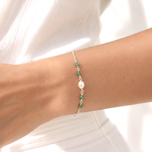 May include: A delicate silver chain bracelet featuring a single white oval pearl centred between four small, round, faceted green gemstone beads. The bracelet is constructed with thin silver links and secures around the wrist with a simple clasp.
