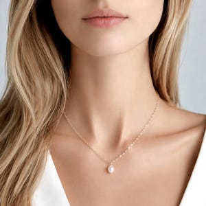 May include: A delicate gold chain necklace featuring a single, small, white, oval-shaped pearl pendant centred on the collarbone. The necklace is constructed with fine, polished gold links that catch the light, creating a subtle and refined appearance.