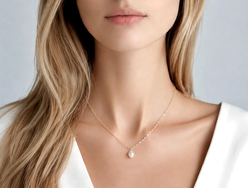 May include: A delicate gold chain necklace featuring a single, small, white, oval-shaped pearl pendant centred on the collarbone. The necklace is constructed with fine, polished gold links that catch the light, creating a subtle and refined appearance.