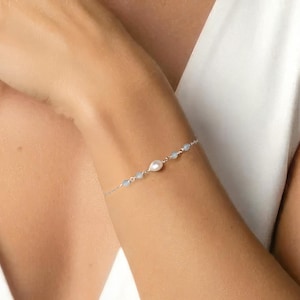May include: A delicate silver bracelet with a central pearl and small, light blue beads. The bracelet is worn on an arm, against a white background.