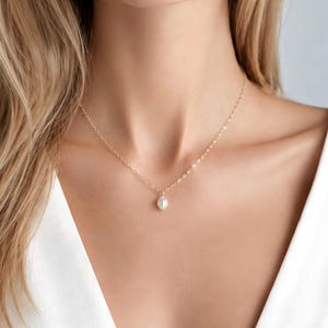 May include: A delicate gold chain necklace featuring a single, small, white oval-shaped pearl pendant. The necklace rests against the neck, showcasing a thin, polished metal link chain that connects to the centred, smooth, lustrous pearl drop.