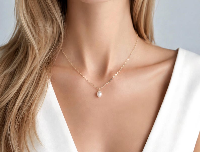 May include: A delicate gold chain necklace featuring a single, small, white oval-shaped pearl pendant. The necklace rests against the neck, showcasing a thin, polished metal link chain that connects to the centred, smooth, lustrous pearl drop.