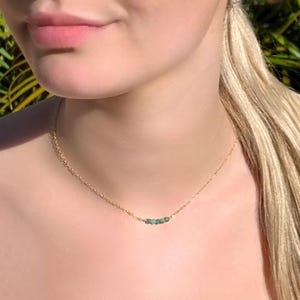 May include: A delicate gold chain necklace featuring a small, horizontal row of tiny, raw, green emerald-coloured gemstone beads centred on the chain. The necklace rests against the neck, showcasing a simple, minimalist metal link design.