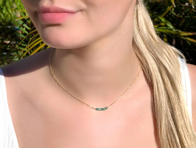 May include: A delicate gold chain necklace featuring a small, horizontal row of tiny, raw, green emerald-coloured gemstone beads centred on the chain. The necklace rests against the neck, showcasing a simple, minimalist metal link design.