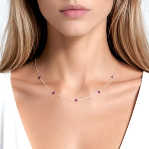 May include: A delicate silver chain necklace featuring five small, evenly spaced, round magenta-colored beads. The necklace rests against the neck, showcasing a minimalist design with a thin metallic link chain and vibrant, polished circular accents.