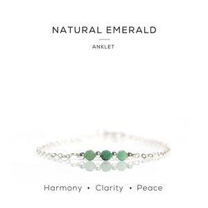 May include: A silver anklet with three natural emerald beads. The text "NATURAL EMERALD ANKLET" is above the anklet. The words "Harmony • Clarity • Peace" are written below the anklet. The anklet is a delicate piece of jewellery.