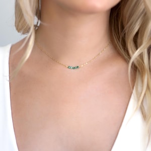 May include: A delicate gold chain necklace featuring three small, round, dark green beads centred on the strand. The necklace rests against the neck, showcasing a thin, polished metal link chain that connects to the trio of vibrant green stones.