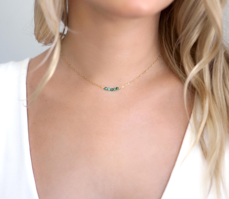 May include: A delicate gold chain necklace featuring three small, round, dark green beads centred on the strand. The necklace rests against the neck, showcasing a thin, polished metal link chain that connects to the trio of vibrant green stones.