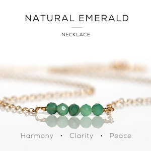 May include: A gold chain necklace featuring five small, faceted green emerald beads arranged in a horizontal row. The text NATURAL EMERALD NECKLACE appears at the top, and the words Harmony, Clarity, and Peace are printed at the bottom.