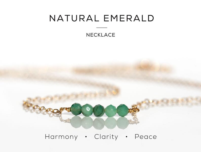 May include: A gold chain necklace featuring five small, faceted green emerald beads arranged in a horizontal row. The text NATURAL EMERALD NECKLACE appears at the top, and the words Harmony, Clarity, and Peace are printed at the bottom.