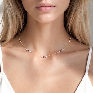 May include: A delicate gold chain necklace featuring a series of small, white, oval-shaped freshwater pearls interspersed with tiny, round, deep pink rubies. The necklace rests against the neck, showcasing a minimalist design with evenly spaced beads.