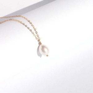 May include: A delicate gold chain necklace featuring a single, smooth, teardrop-shaped white pearl pendant. The pearl is securely attached to the fine metal link chain, which displays a warm, reflective metallic lustre against the bright surface.