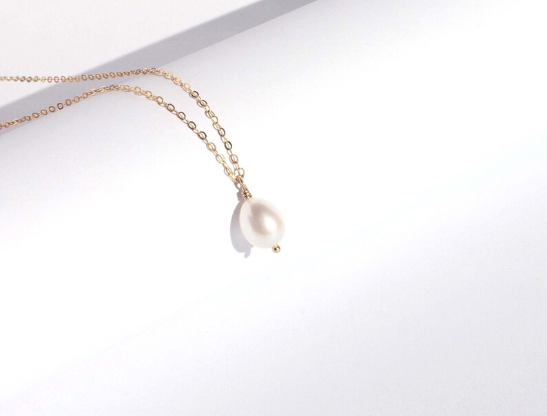 May include: A delicate gold chain necklace featuring a single, smooth, teardrop-shaped white pearl pendant. The pearl is securely attached to the fine metal link chain, which displays a warm, reflective metallic lustre against the bright surface.