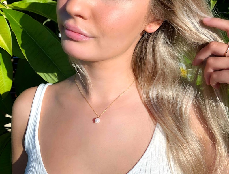 May include: A delicate gold chain necklace featuring a single, small, white, oval-shaped pearl pendant. The necklace rests against the neck, showcasing a thin, polished gold metal link chain that holds the pearl securely in the centre position.