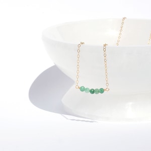 May include: A delicate gold chain necklace featuring a horizontal row of five small, faceted, round green gemstone beads centred on the chain. The necklace is draped over the edge of a smooth, white, translucent, pedestal-style bowl.