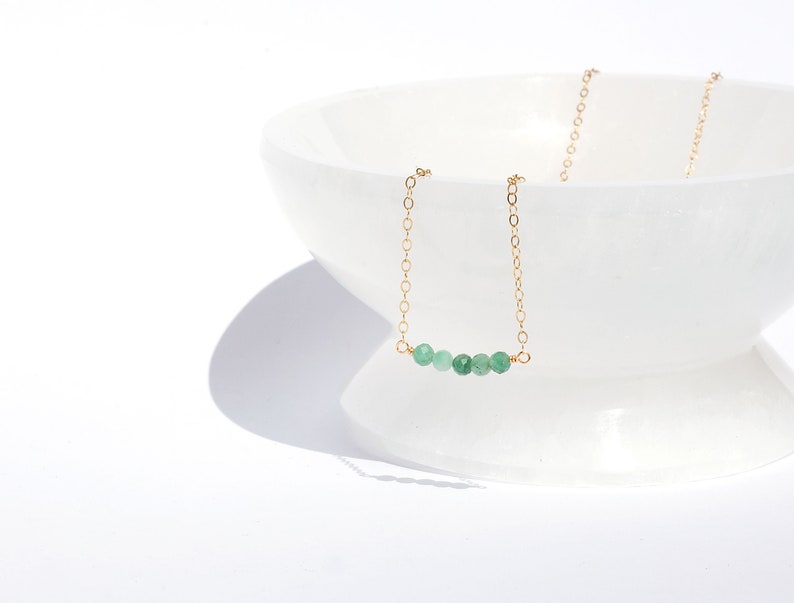 May include: A delicate gold chain necklace featuring a horizontal row of five small, faceted, round green gemstone beads centred on the chain. The necklace is draped over the edge of a smooth, white, translucent, pedestal-style bowl.