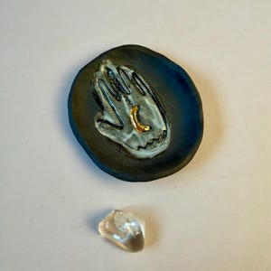 May include: A dark blue ceramic dish with a hand design in white and gold, and a clear quartz crystal. The dish has a handprint design with a gold crescent moon. The quartz crystal is clear and smooth.