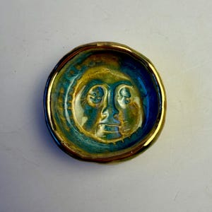 Op de afbeelding: Een kleine, ronde keramische schaal met een gezichtsontwerp. De schaal heeft een blauw en goud glazuur met een gouden rand. Het gezicht is een eenvoudige, gestileerde weergave met ogen, neus en mond.