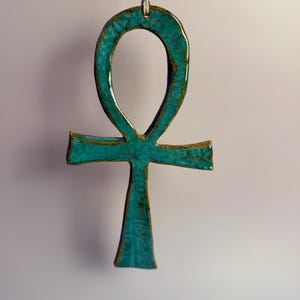 Może przedstawiać: Turkusowy wisiorek Ankh z teksturowaną powierzchnią i złotymi akcentami, zawieszony na cienkim, białawym sznurku. Starożytny egipski symbol jest prezentowany na miękkim, neutralnym tle.