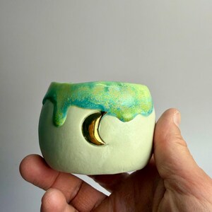 May include: A small, round, ceramic planter in a pale green color with a gold crescent moon cutout. The top edge has a dripping glaze in shades of green and blue. The planter is held in a hand.