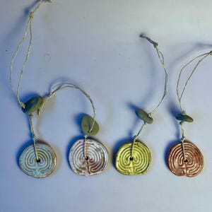Pode incluir: Quatro pingentes de cerâmica feitos à mão com um design de labirinto. Cada pingente tem uma cor diferente: azul claro, branco e rosa, amarelo e marrom avermelhado. Eles são amarrados em barbante natural com uma pequena conta de pedra.