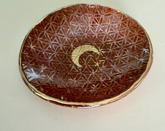May include: A round, shallow ceramic dish with a glossy, reddish-brown finish featuring an embossed geometric flower of life pattern. A gold-colored crescent moon is centered in the bowl, and the rim is finished with a metallic gold-colored edge.