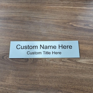 May include: A rectangular, brushed metal nameplate with the text "Custom Name Here" above "Custom Title Here" in black lettering. The nameplate is set against a brown wooden surface.