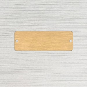 May include: A gold-colored metal tag with two holes for hanging. The tag has a brushed finish.
