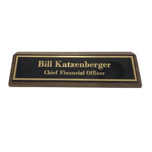 May include: A rectangular desk nameplate with a dark brown wooden base and a black center. The nameplate features the name "Bill Katzenberger" and the title "Chief Financial Officer" in gold lettering. The nameplate has a gold border.