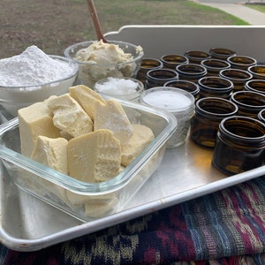 Pu&ograve; includere: Un vassoio di metallo con vari ingredienti per la preparazione di burro corpo fatto in casa, tra cui burro di karit&egrave;, olio di cocco e oli essenziali. Gli ingredienti sono in barattoli e ciotole di vetro, e ci sono anche piccoli barattoli di vetro per confezionare il prodotto finito.