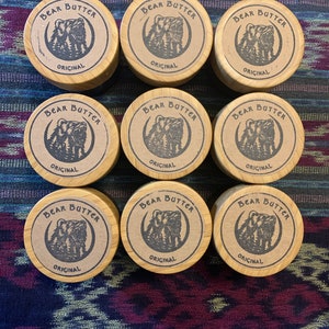 Pu&ograve; includere: Nove contenitori rotondi in legno con coperchi marroni. Ogni coperchio ha un'illustrazione in bianco e nero di un orso con il testo "Bear Butter Original" stampato su di esso.