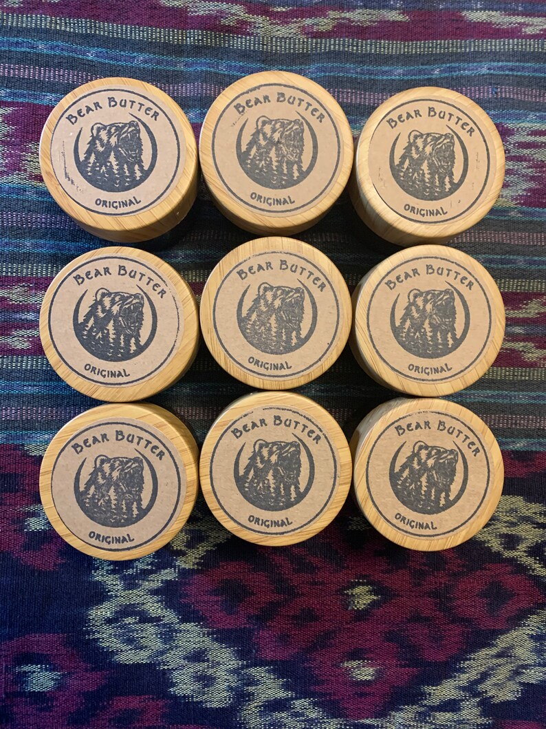 Pu&ograve; includere: Nove contenitori rotondi in legno con coperchi marroni. Ogni coperchio ha un'illustrazione in bianco e nero di un orso con il testo "Bear Butter Original" stampato su di esso.